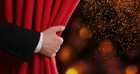 A hand is seen pulling back a red theatre curtain.