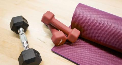 A rolled-up burgundy yoga mat and three free weights sit on a wooden fitness studio floor.