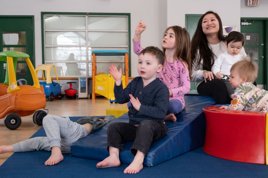 Singing, while learning to listen and participate in class activities at circle time. Five children, ages 1 to 5 sing songs while sitting on play equipment in a play room.