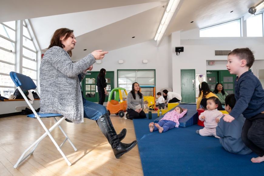Circle time with Amy An instructor sitting on a chair leads young children sitting on a mat in a song in a play room.