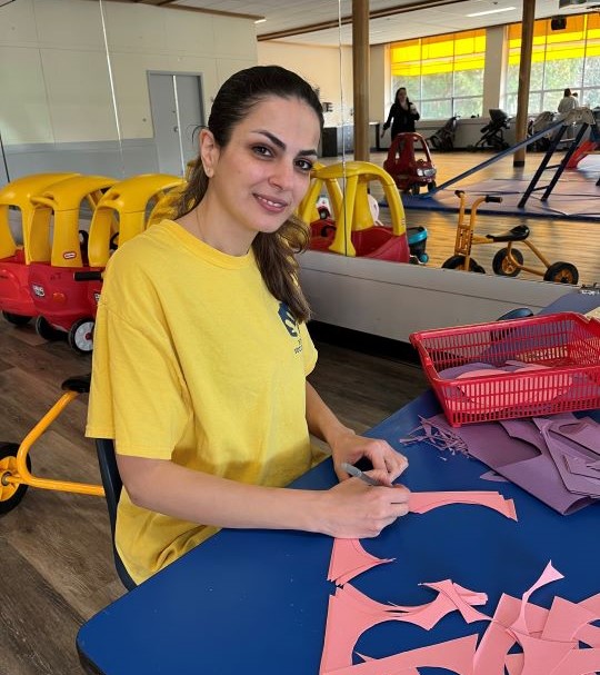 Toddler & Me volunteer, Mina prepares supplies for a fun craft activity. A woman in a yellow t-shirt cuts out pink paper scraps for a Toddler & Me craft activity.