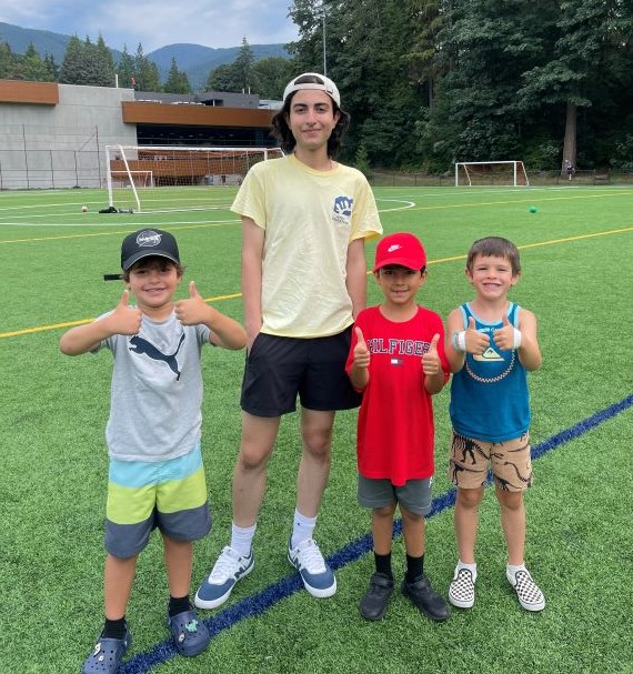 Our camp volunteers help kids get the most out of their summer vacation! A teen boy in a volunteer's yellow t-shirt stands on a playing field with three younger boys giving thumbs up.