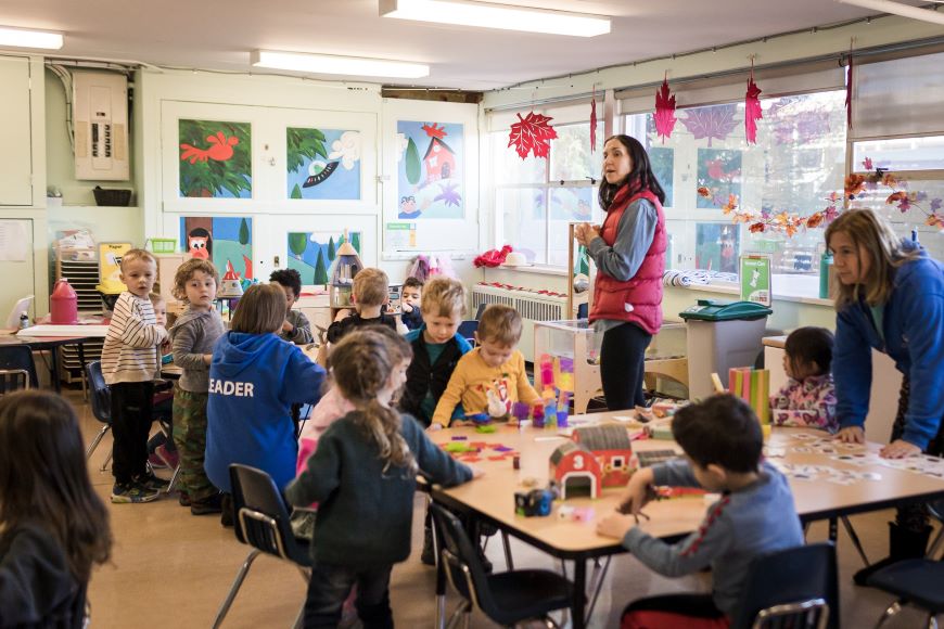 The teachers at the Lonsdale Preschool watch as the children explore their interests during free play. Three preschool teachers play with groups of children at classroom tables exploring a variety of toys and games.
