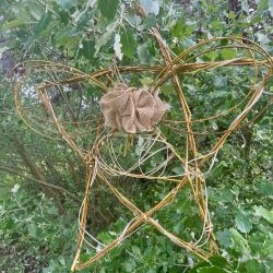 Stars of the Neighbourhood A woven willow star hangs from a leafy tree branch in a park.