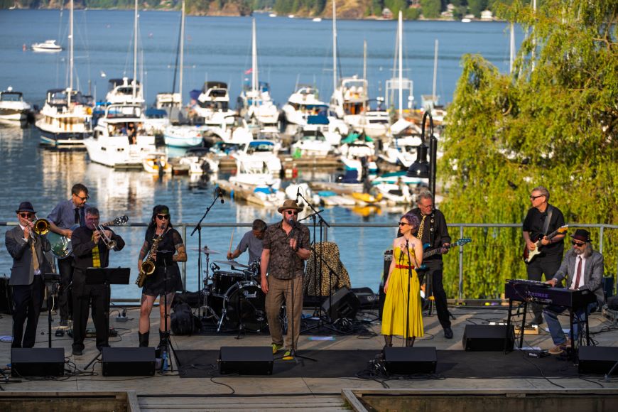 A band performs on a stage in Panorama Park, in front of Deep Cove. 
