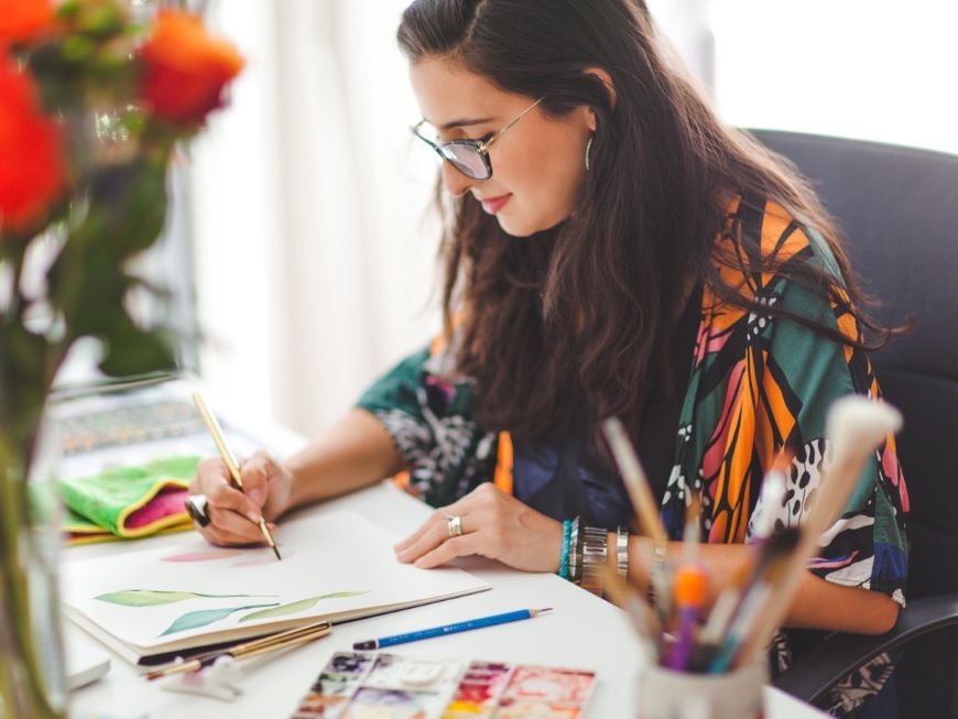 An artist sits at a table, painting a watercolour. 