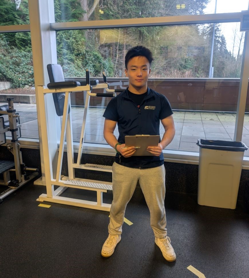 A practicum student holding a clip board and wearing an NVRC vest works in the Delbrook Fitness Centre.