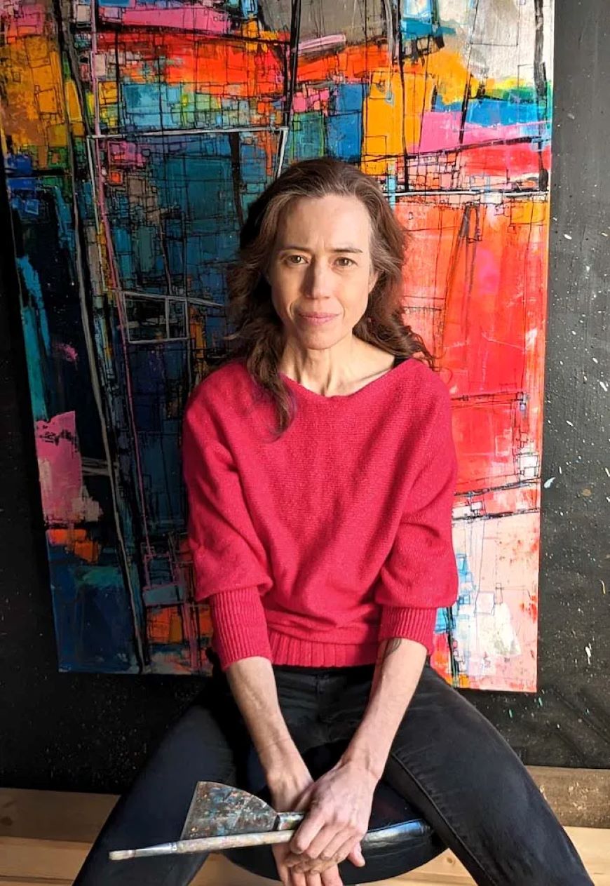 An artist in a red sweater sits on a stool holding a pant brush and pallet knife, in front of a large, colourful abstract painting. 