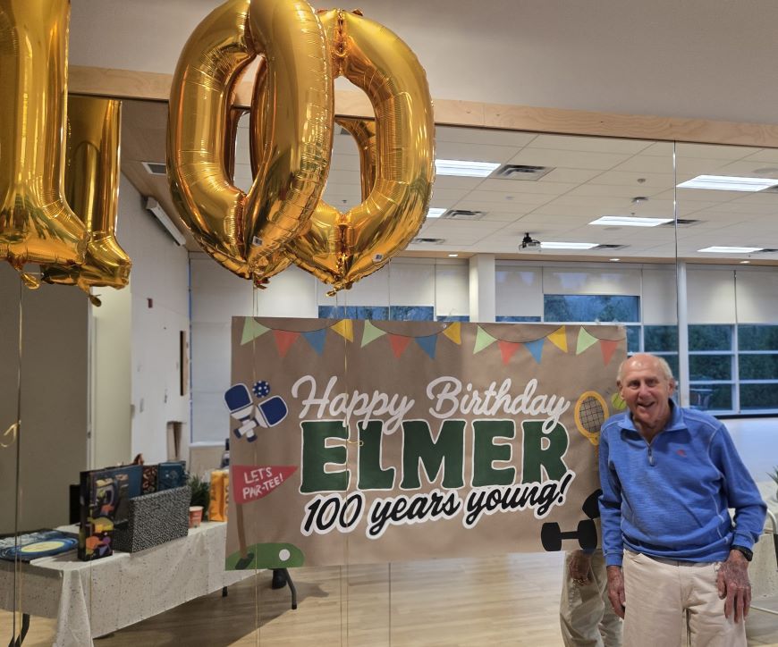 A gentleman stands in front of Happy 100th Birthday banner.