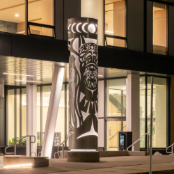 Illuminated steel sculpture featuring a Sḵwx̱wú7mesh Thunderbird, in front of a modern residential building.