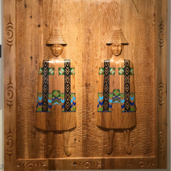 This large carved and painted cedar panel depicts the twin daughters of the Sḵwx̱wú7mesh (Squamish) Chief standing between the Twin Sisters mountain peaks in noble Coast Salish woven robes. 