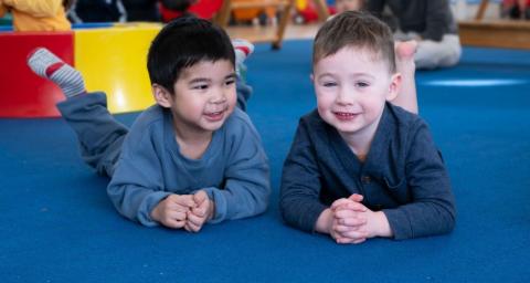 Two smiling toddler boys lie on a mat in a play room.