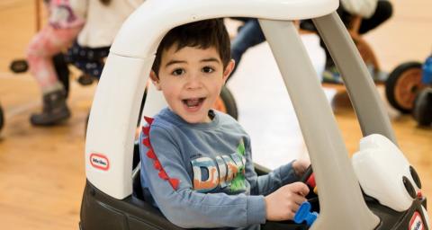 A preschool-aged boy smiles while riding in a toy car in a gymnasium with other children on bikes in the background. 
