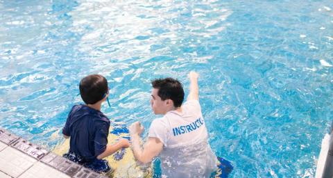 A young boy stands in the pool next to a swim instructor.