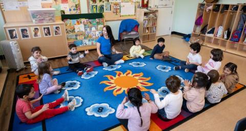 A preschool teacher leads a circle of children seated cross-legged on a colourful carpet in a song. 