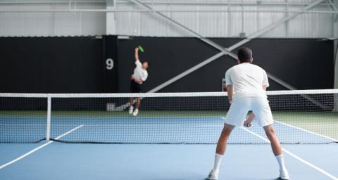 Two men play tennis on a court. The man in the background is jumping to serve, the man in the foreground is leaning forward in a ready position. 