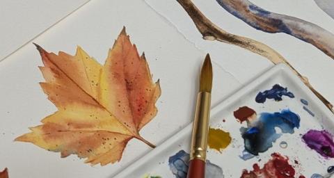 An artist's watercolour works in-progress on a table, including a palette and brush, colour swatches, and paintings of a leaf and branches are visible. 