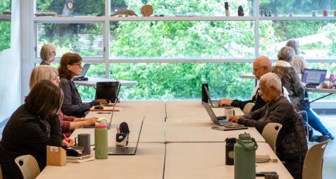 A group of people work on laptops at a large table in a room with big windows. 