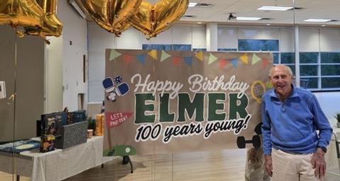 A gentleman stands in front of Happy 100th Birthday banner.