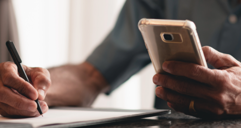 A man holds a phone while writing a list with the other.