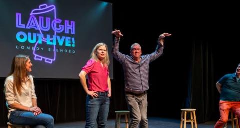 Four comedians on a stage. Two sitting on stools, two standing. One has raised arms. 