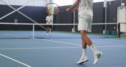 A man hits a tennis ball towards an opponent on an indoor tennis court. 