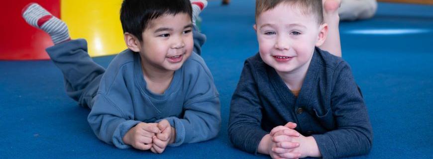 Two smiling toddler boys lie on a mat in a play room. 