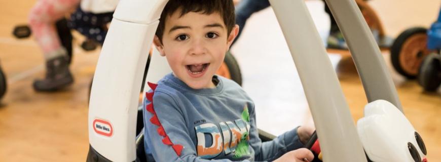 A preschool-aged boy smiles while riding in a toy car in a gymnasium with other children on bikes in the background. 
