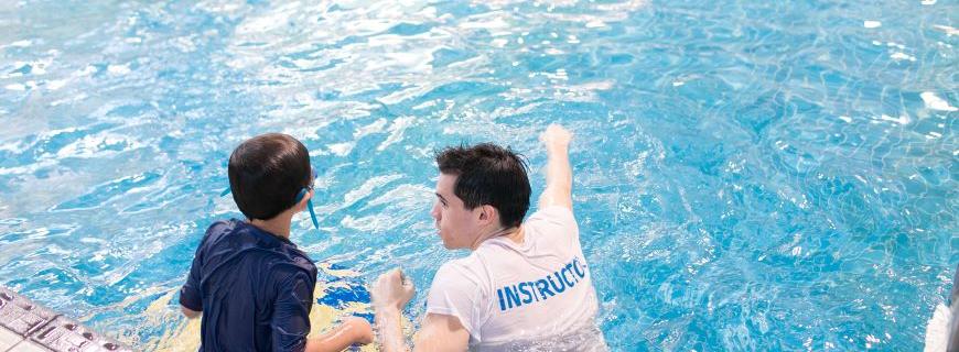 A young boy stands in the pool next to a swim instructor. 