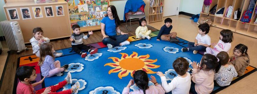 A preschool teacher leads a circle of children seated cross-legged on a colourful carpet in a song. 