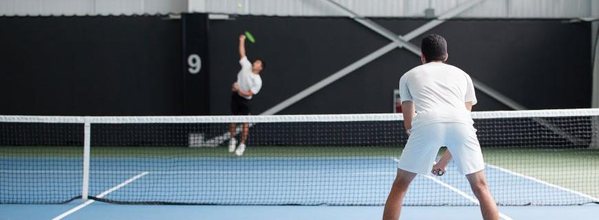 Two men play tennis on a court. The man in the background is jumping to serve, the man in the foreground is leaning forward in a ready position. 