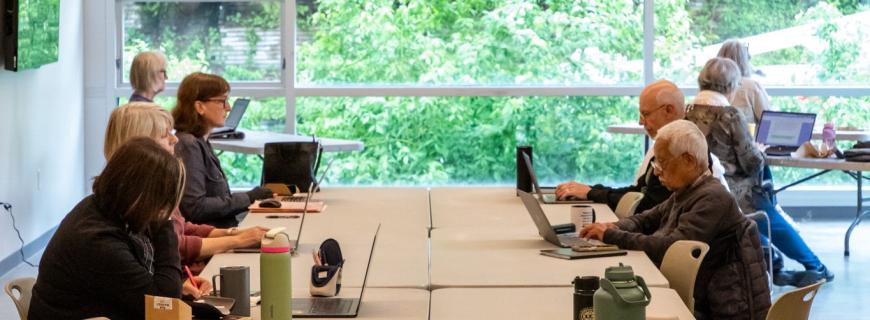 A group of people work on laptops at a large table in a room with big windows. 