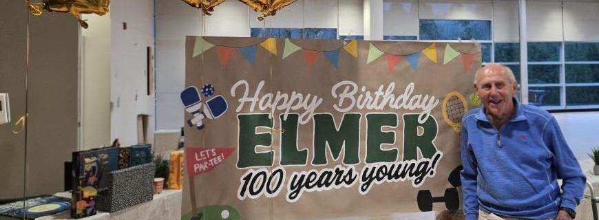 A gentleman stands in front of Happy 100th Birthday banner.