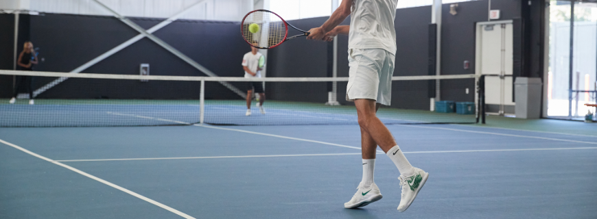 A man hits a tennis ball towards an opponent on an indoor tennis court. 