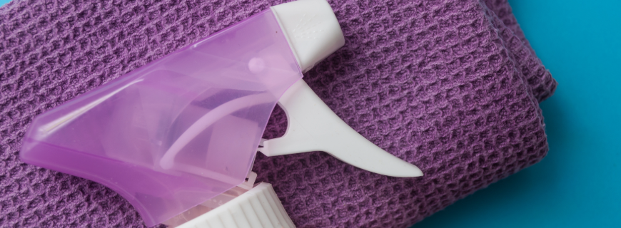 A folded purple cloth towel, with a white and purple spray bottle resting on top. 