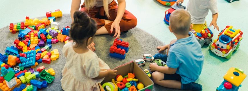 A group of young children sits on the floor with a teacher, playing with toys. 